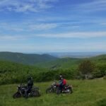 Deux motards avec leurs motos garées sur une colline herbeuse, profitant d'une vue panoramique sur un vaste paysage de montagnes boisées sous un ciel bleu.
